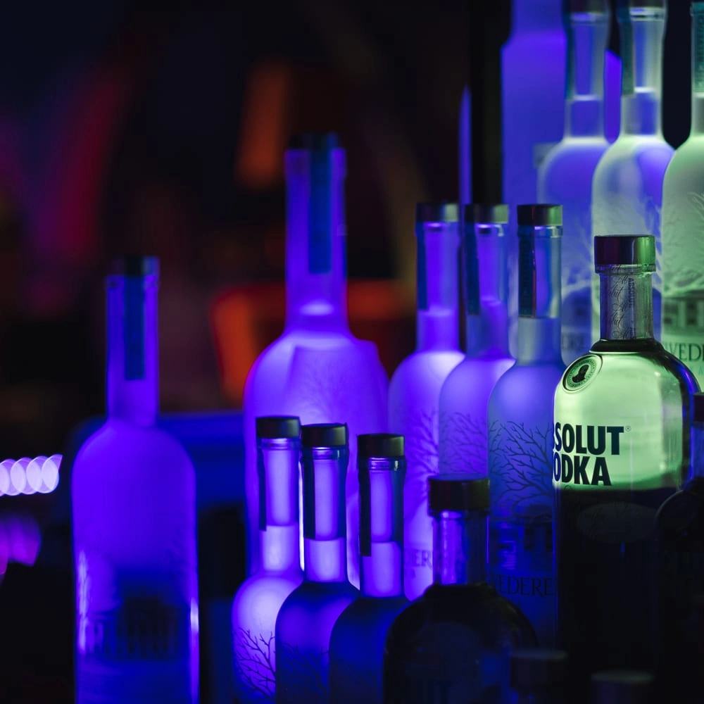 Bouteilles alignées derrière un bar sous des lumières bleues, violettes et vertes.