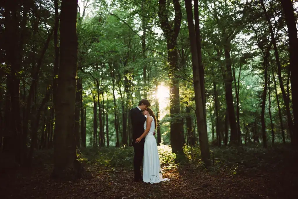 Couple élégamment vêtu s'embrassant dans une forêt traversée par une lumière dorée.