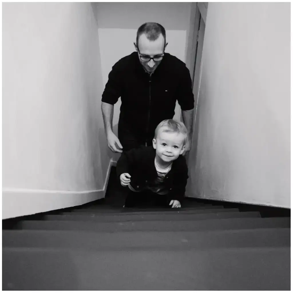 Jeune enfant montant un escalier devant son père pendant une séance photo de famille en noir et blanc.