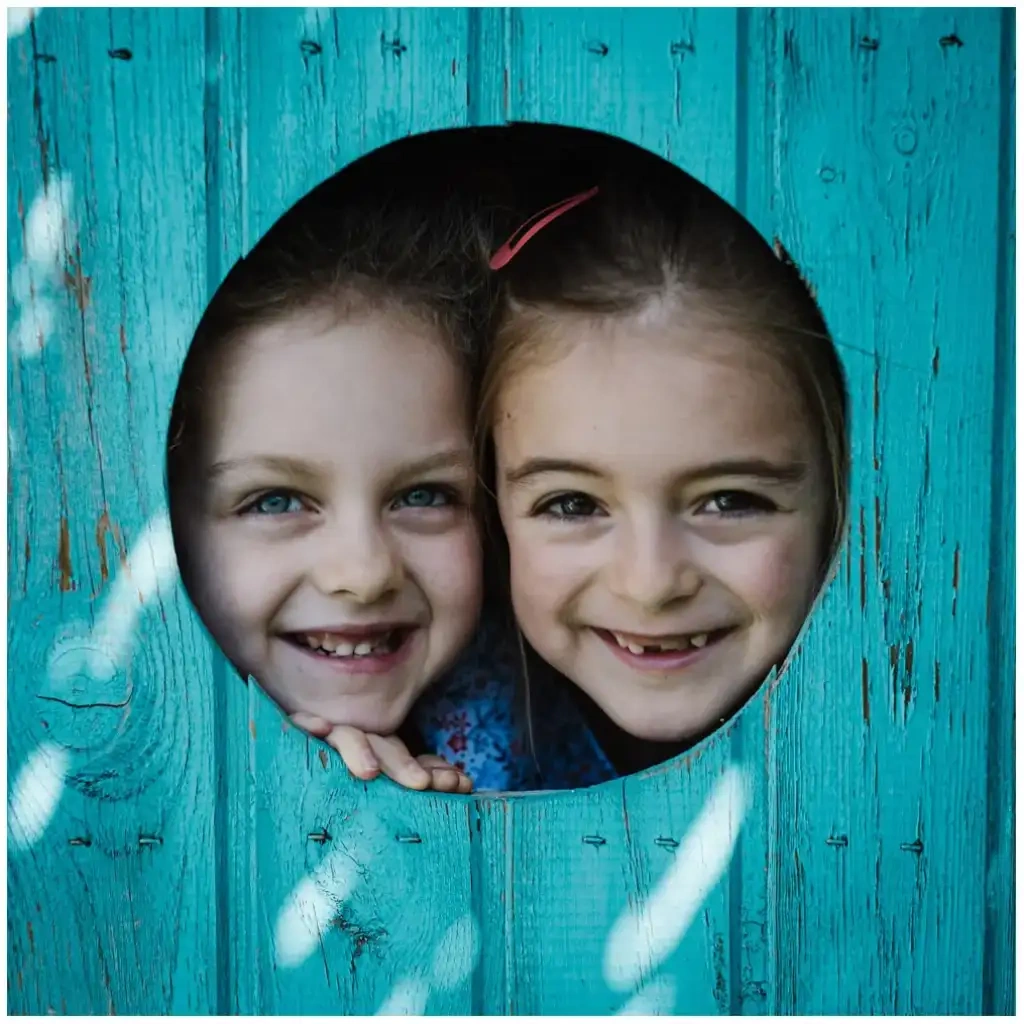 Deux jeunes filles souriantes regardant à travers une ouverture ronde dans une paroi en bois turquoise.