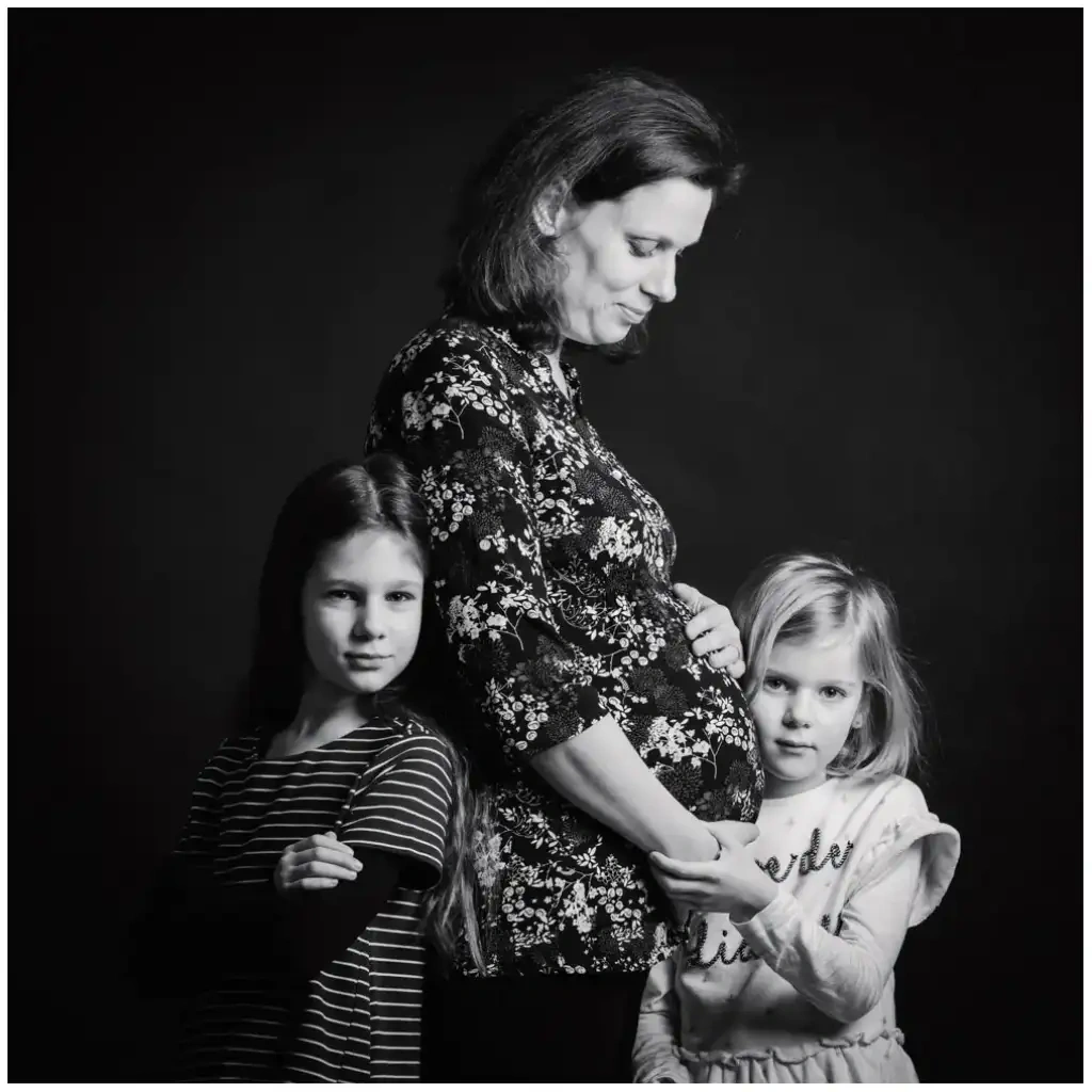 Femme enceinte entourée de deux jeunes filles pendant une séance photo de grossesse en noir et blanc.