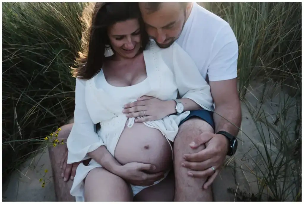 Couple assis dans les dunes autour du ventre de la future mère pendant une séance photo de grossesse.