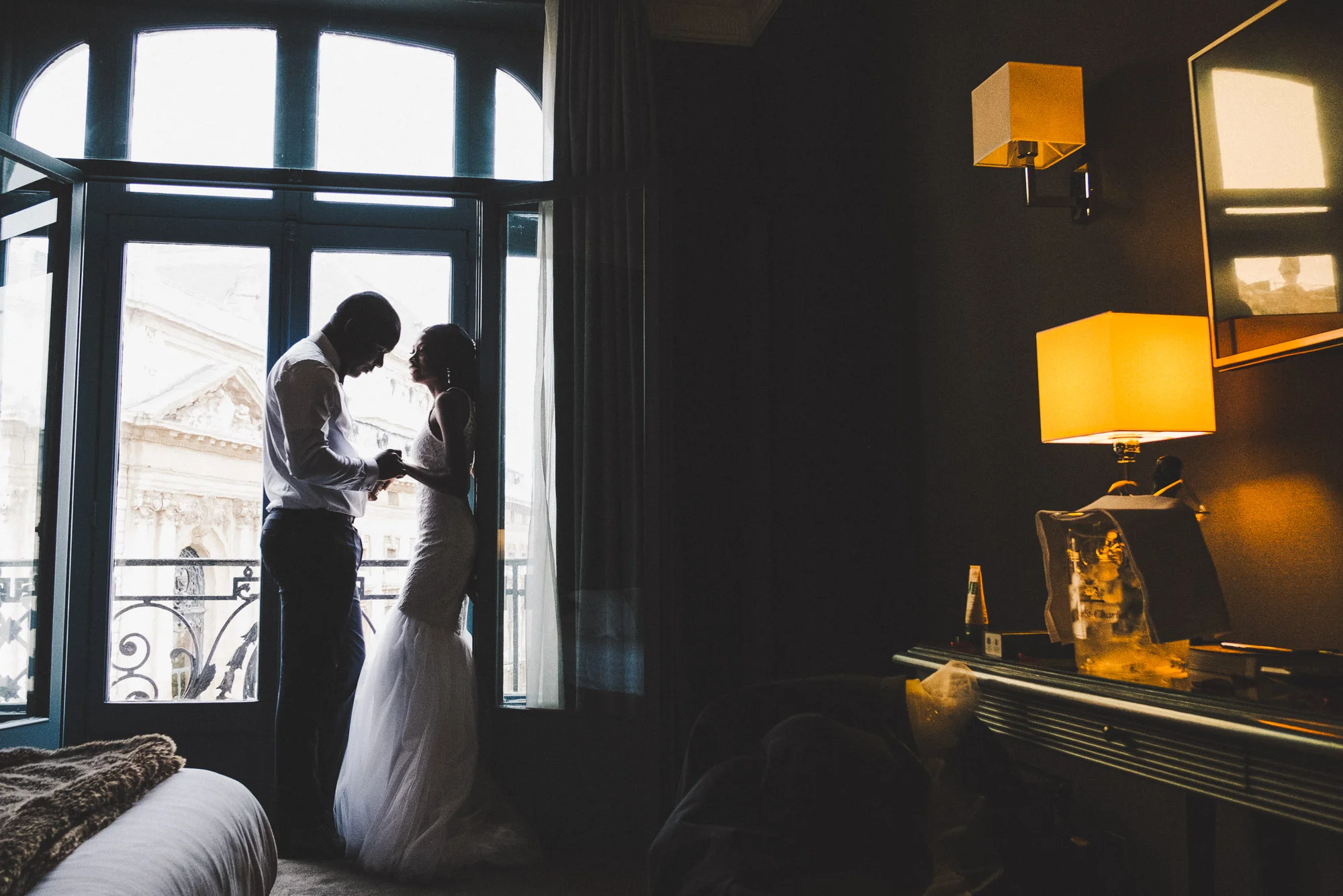 Couple de mariés près d'une fenêtre dans une chambre sombre éclairée par des lampes ambrées.