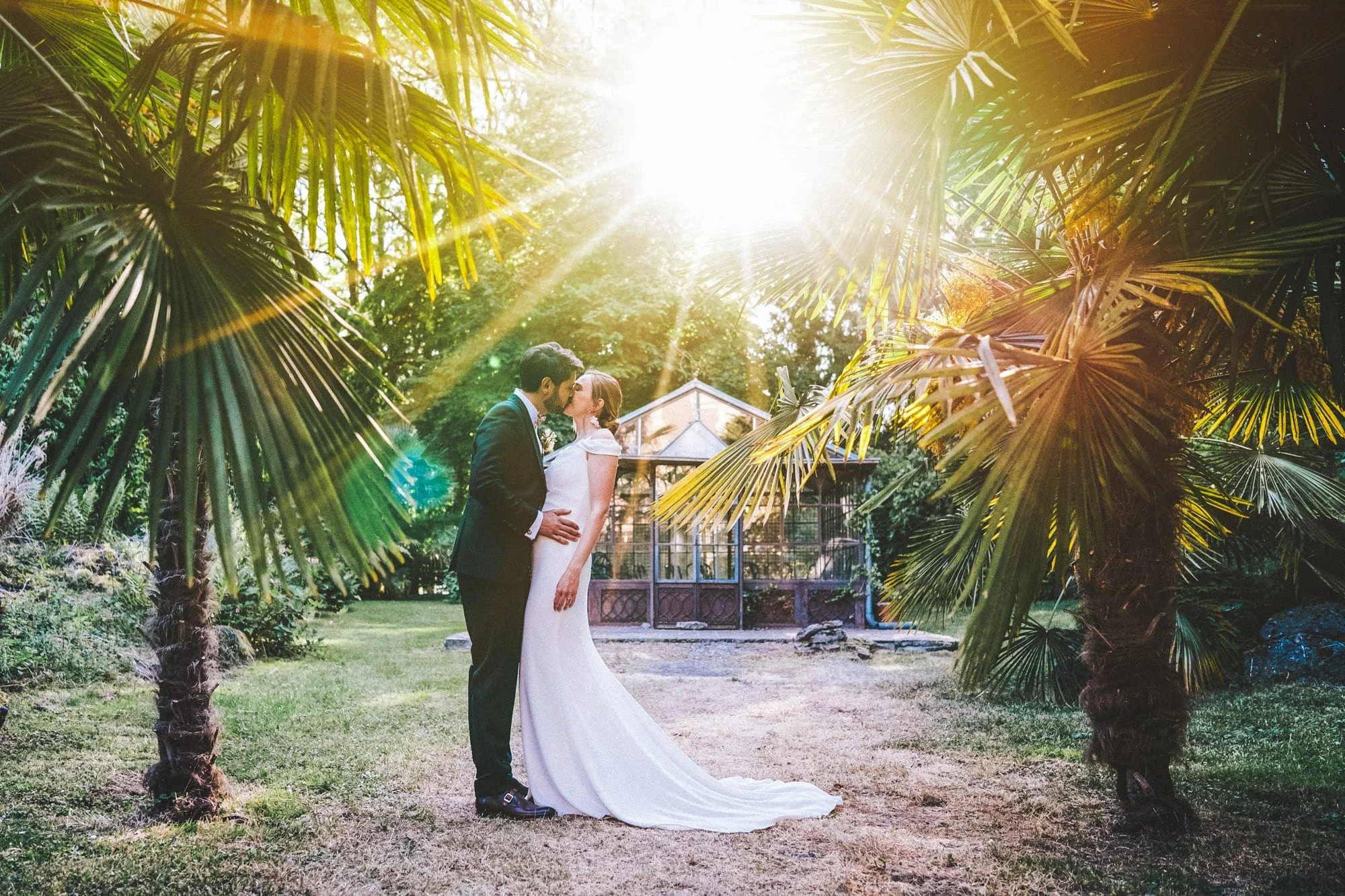 Couple de mariés s'embrassant dans un jardin planté de palmiers sous une lumière dorée.