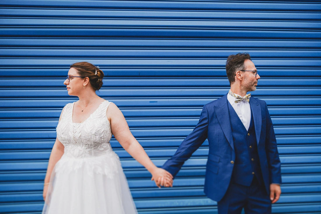 Mariés se tenant par la main devant un rideau métallique bleu lors d'un reportage de mariage.