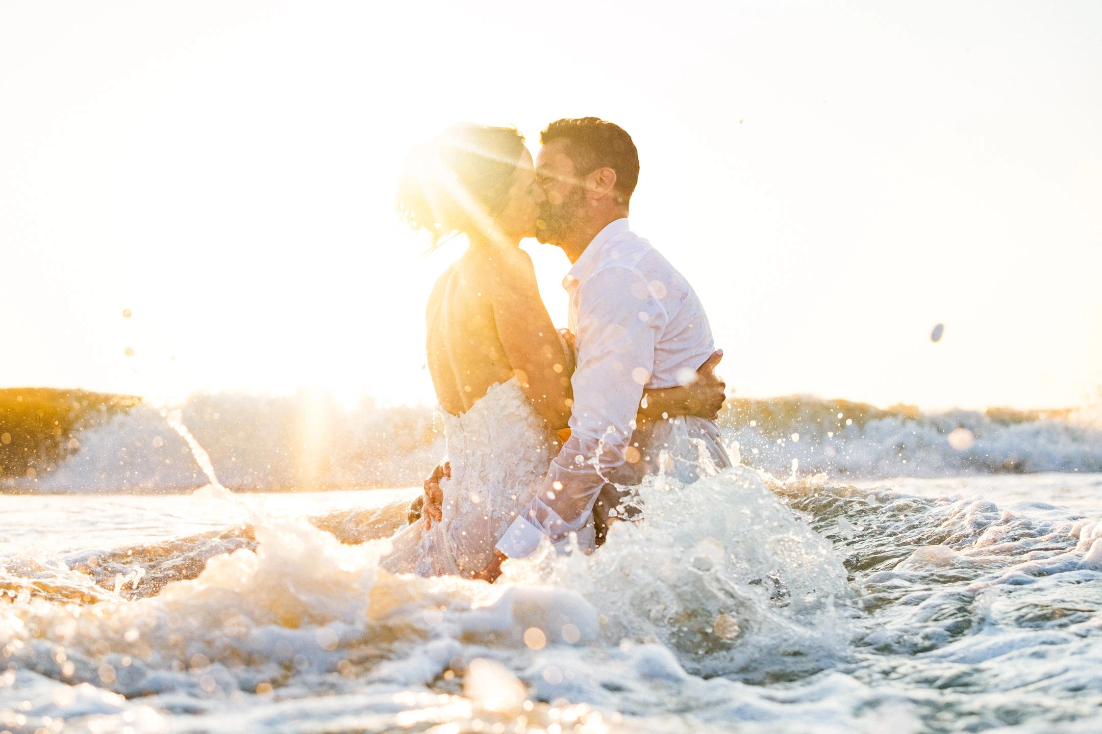 Caroline et Anthony s’embrassant au milieu des vagues dans la lumière du soleil couchant.