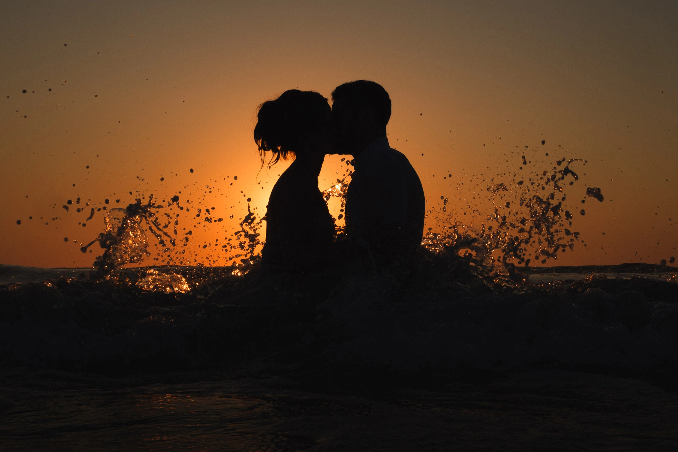 Silhouette de Caroline et Anthony s’embrassant derrière les éclaboussures d’une vague au coucher du soleil.