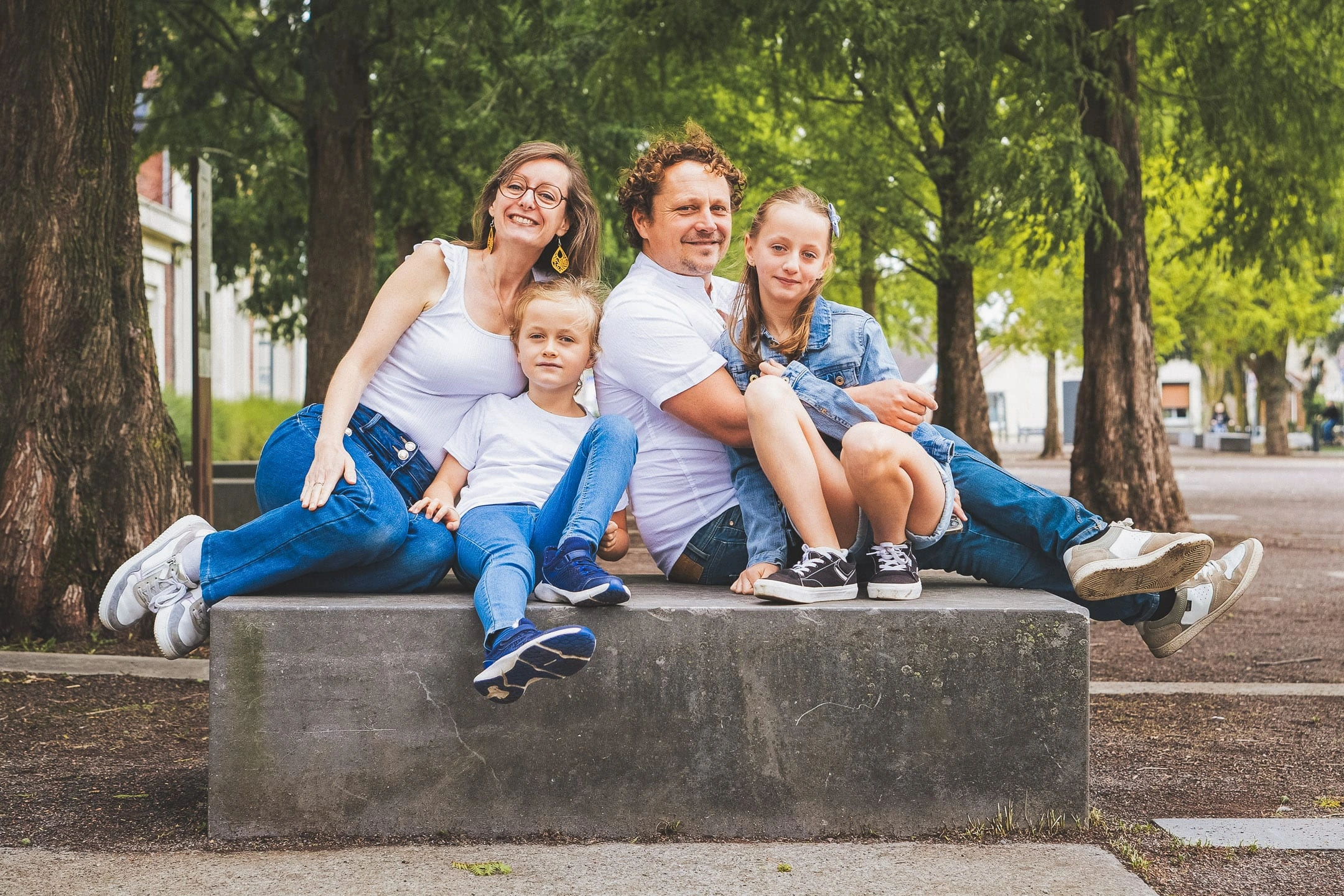 Deux parents et leurs deux filles assis ensemble sur un banc entre des arbres.