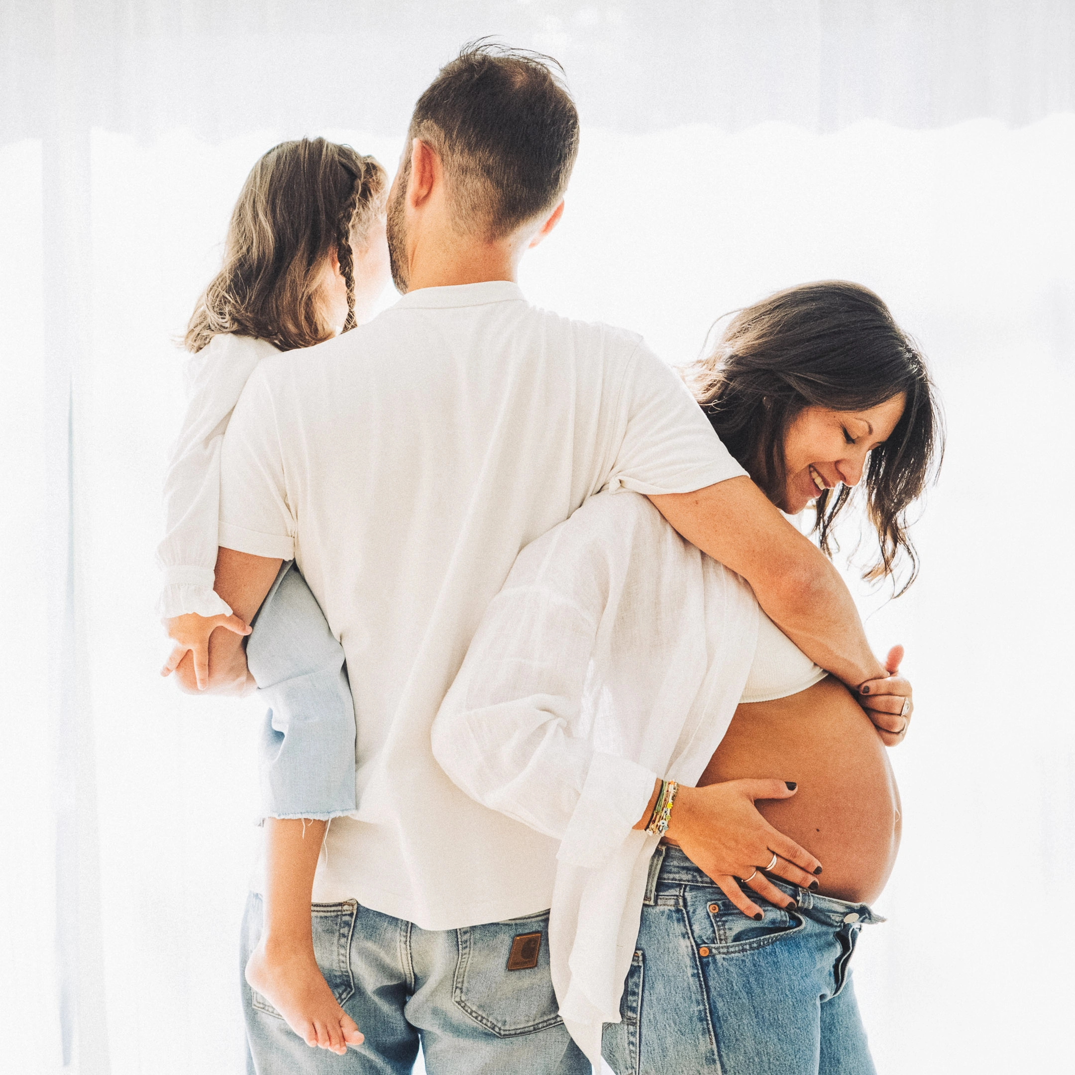 Une future maman souriante, son compagnon et leur fille réunis dans une pièce baignée de lumière.