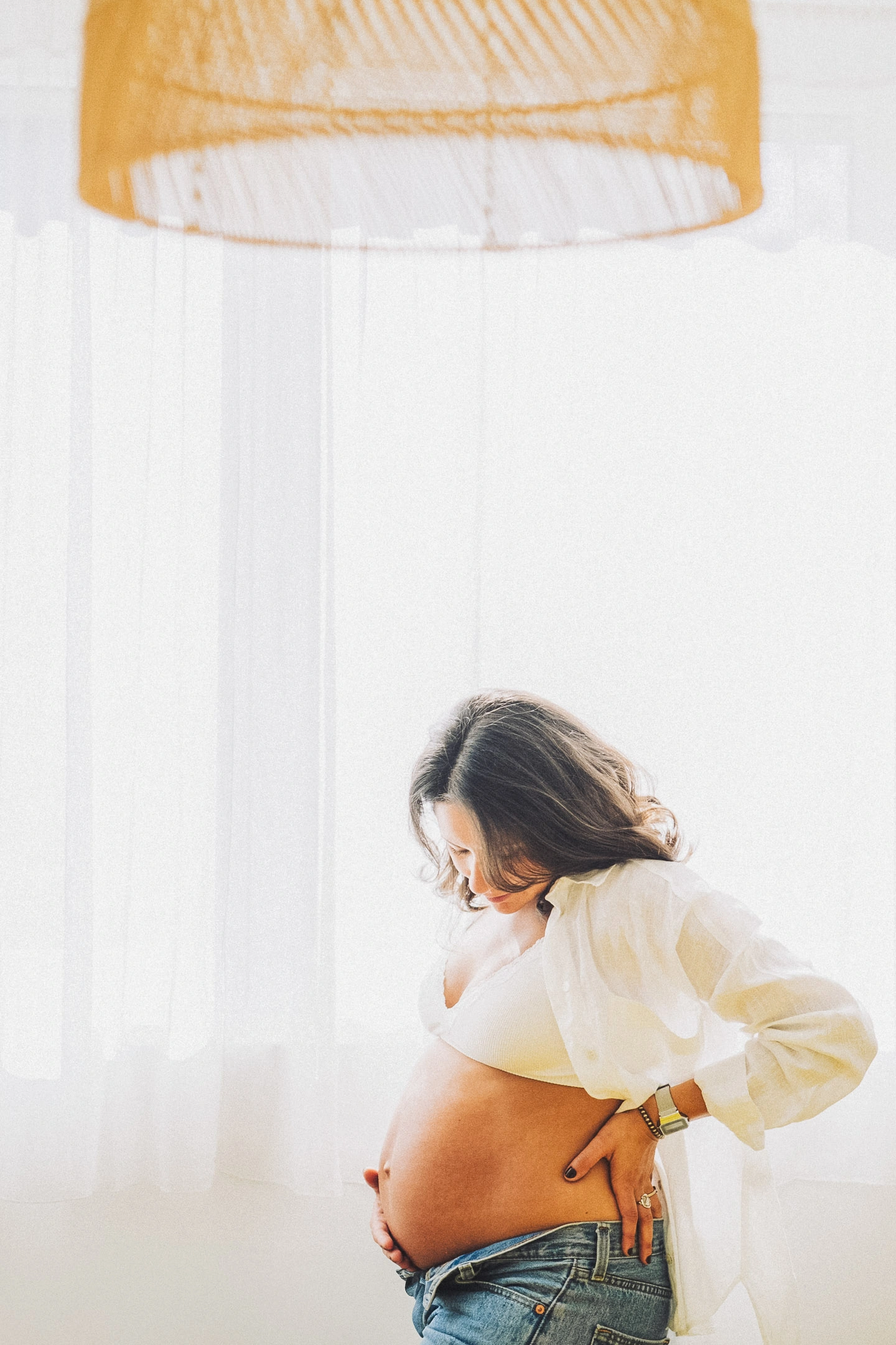 Une future maman en chemise blanche regarde son ventre sous une suspension jaune.