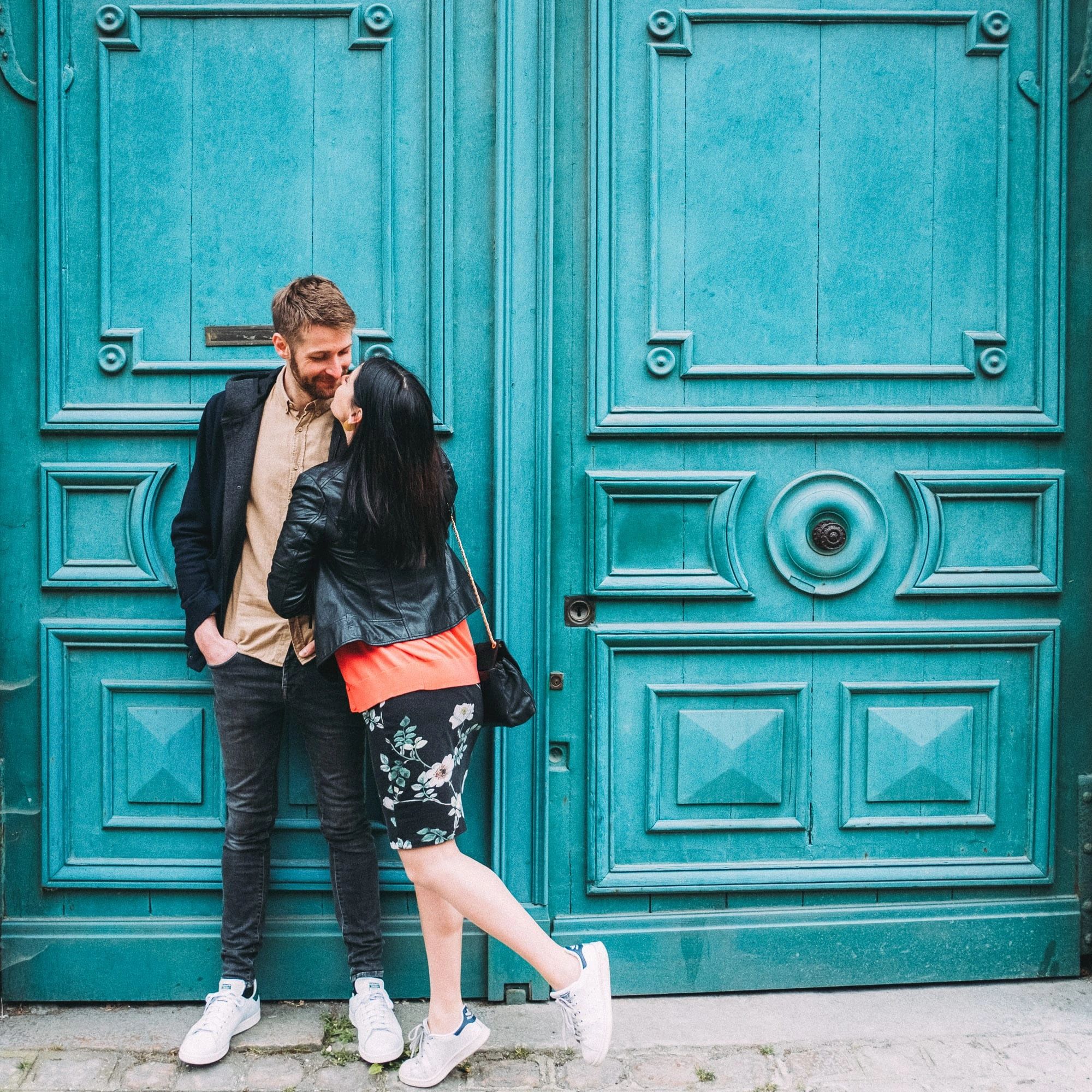 Couple debout devant une grande porte turquoise sculptée, la femme se penchant vers son compagnon.