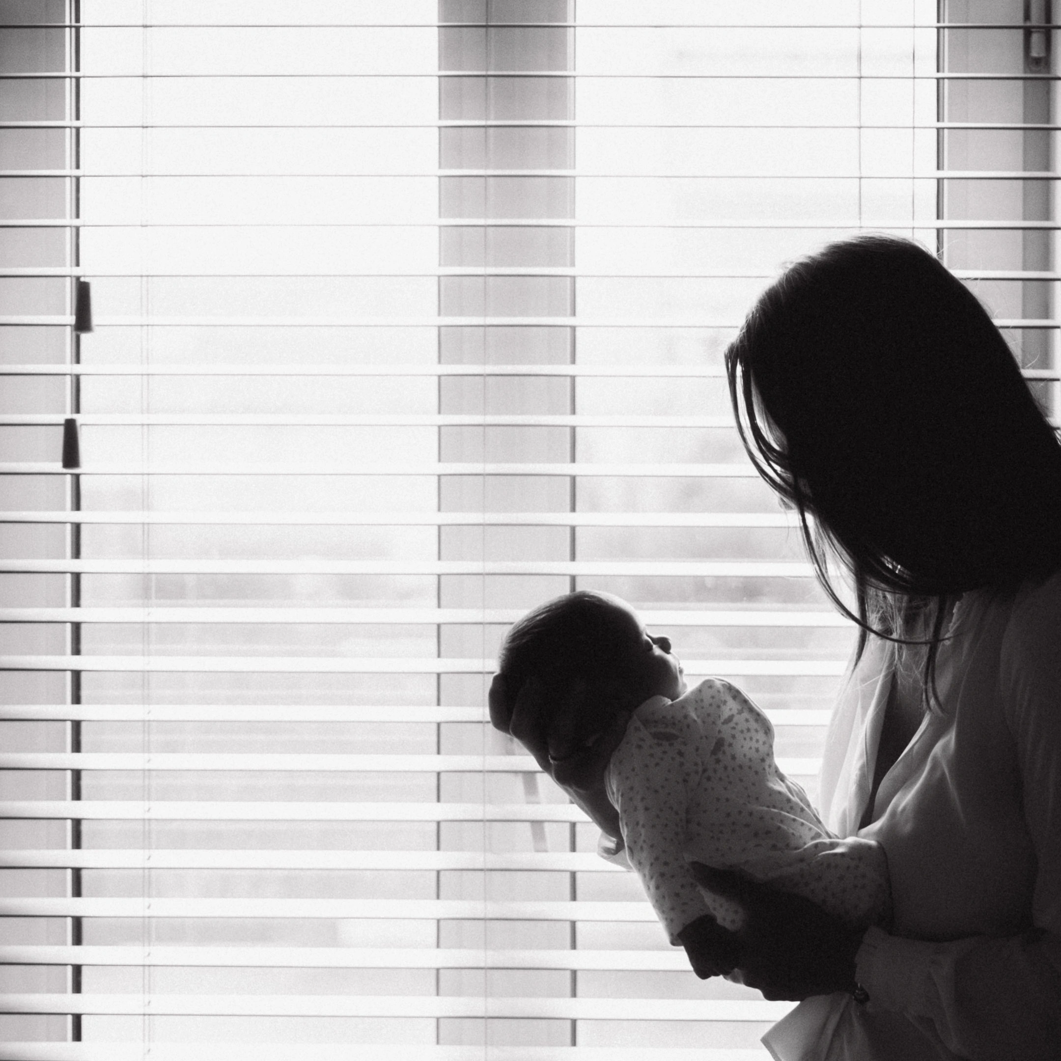 Silhouette en noir et blanc d'une mère tenant son nouveau-né devant une fenêtre à stores.