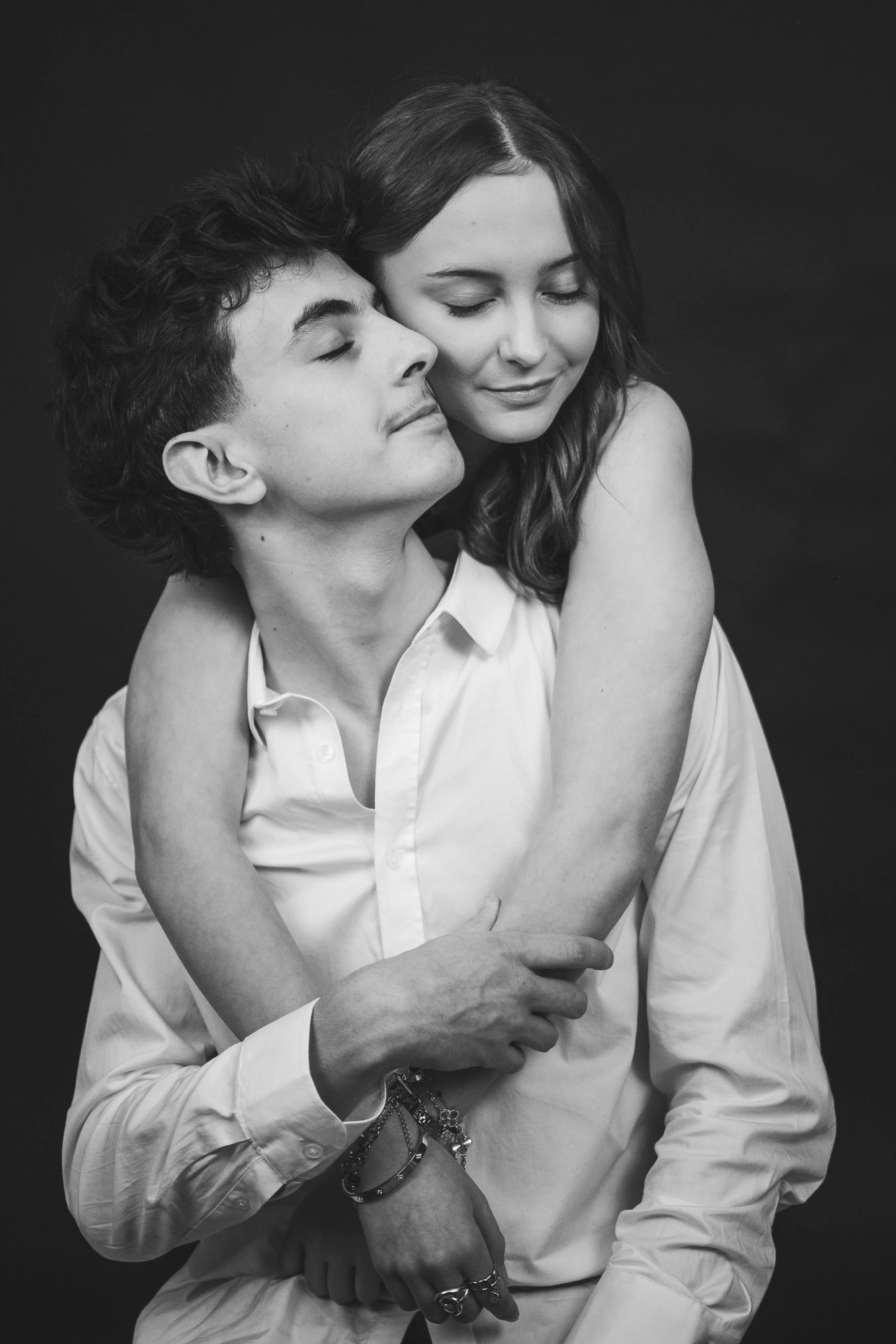 Portrait en noir et blanc d’une femme entourant son compagnon par derrière, les yeux fermés.