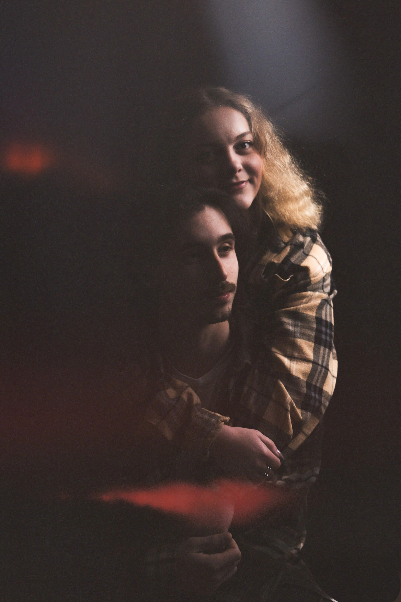Femme en chemise à carreaux entourant son compagnon dans un portrait sombre aux reflets rouges.