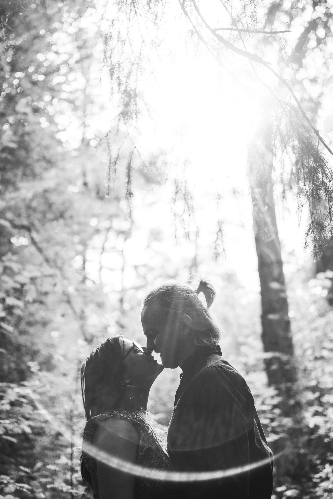 Silhouette en noir et blanc d’un couple rapproché dans une forêt traversée par une lumière vive.
