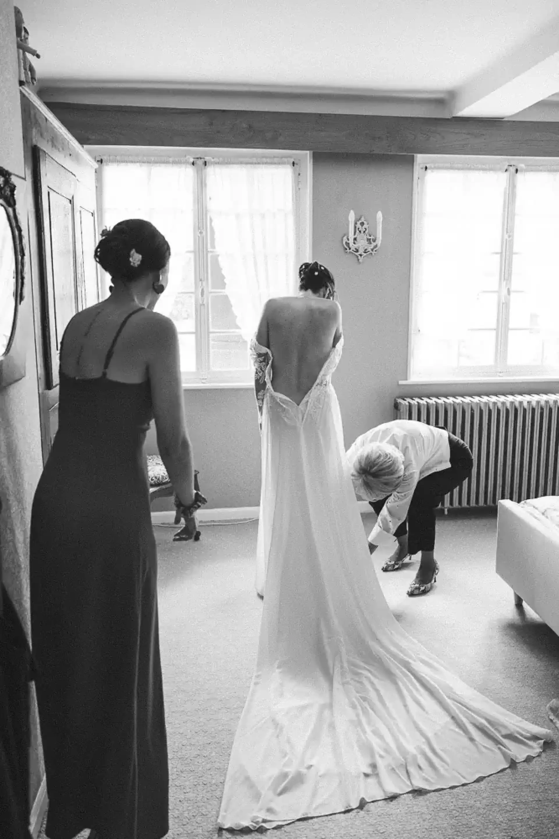 Léa enfilant sa robe avec l'aide de deux proches dans une chambre lumineuse, en noir et blanc.