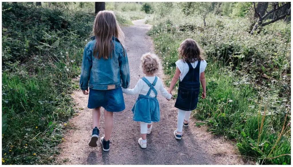 Trois jeunes filles marchant main dans la main sur un sentier entouré de végétation.