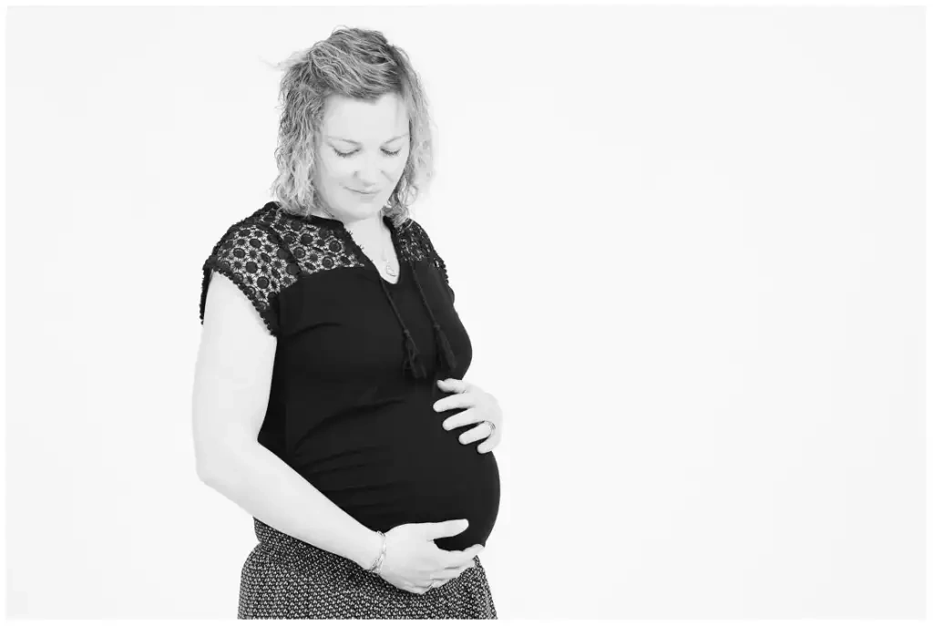 Portrait lumineux en noir et blanc d’une femme enceinte, les mains posées sur son ventre.
