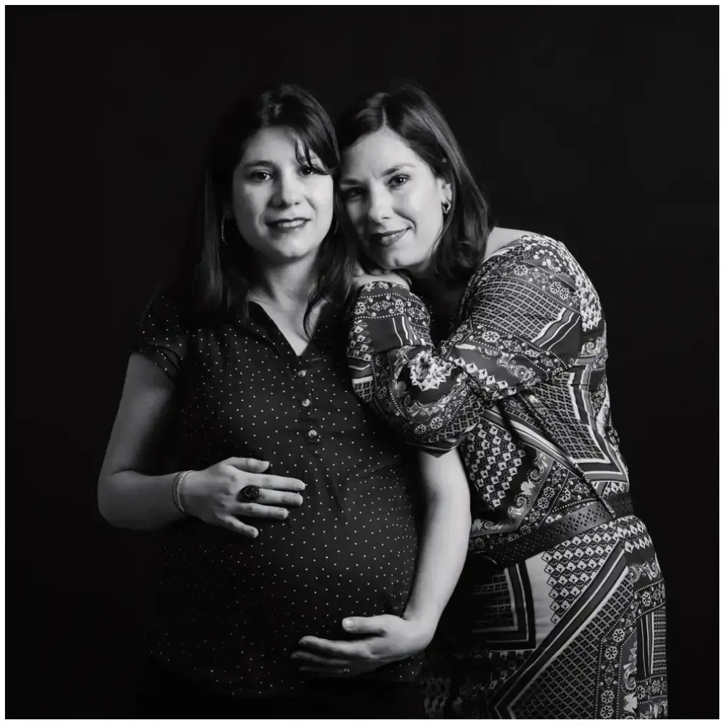 Portrait en noir et blanc de deux amies, dont une femme enceinte, devant un fond sombre.