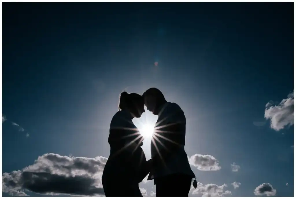 Silhouette d'un couple attendant un enfant, front contre front devant un ciel bleu et le soleil.