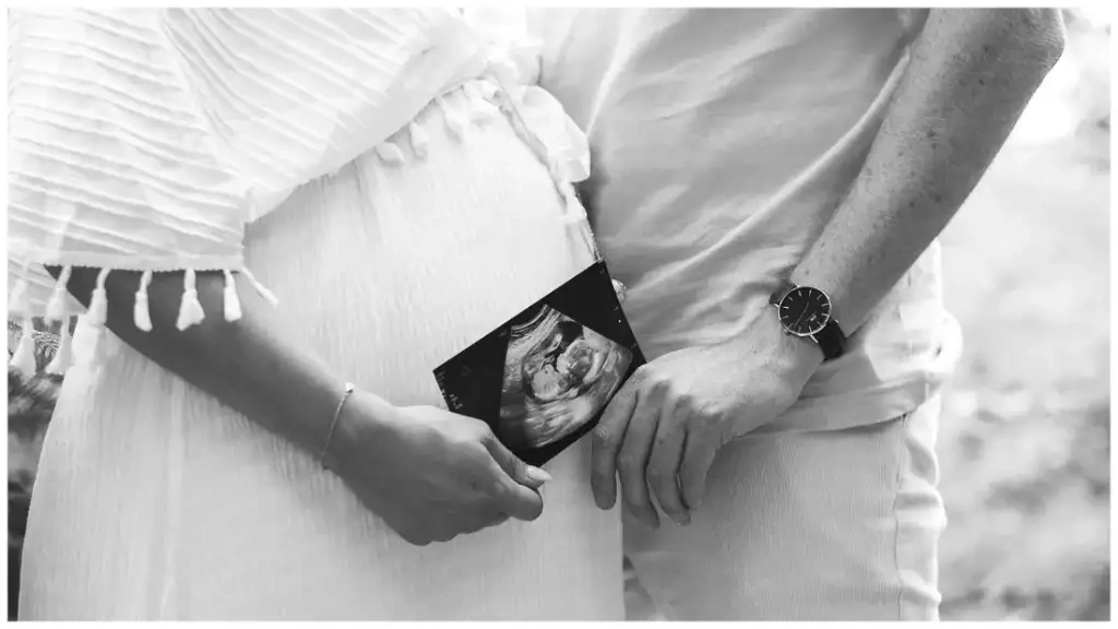 Un couple tient une échographie devant le ventre de grossesse, dans un portrait en noir et blanc.
