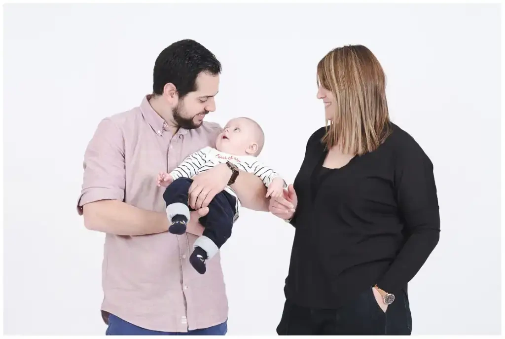 Deux parents regardent leur bébé tenu dans les bras de son père devant un fond clair.