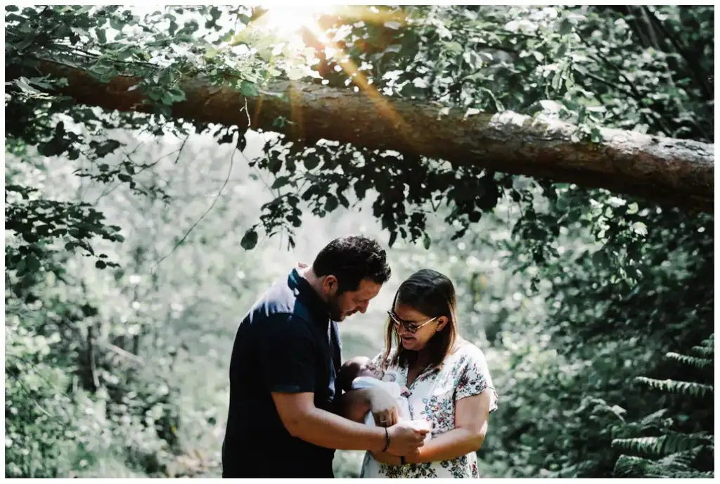 Deux parents regardent leur bébé dans les bras de sa mère au milieu d’une forêt ensoleillée.