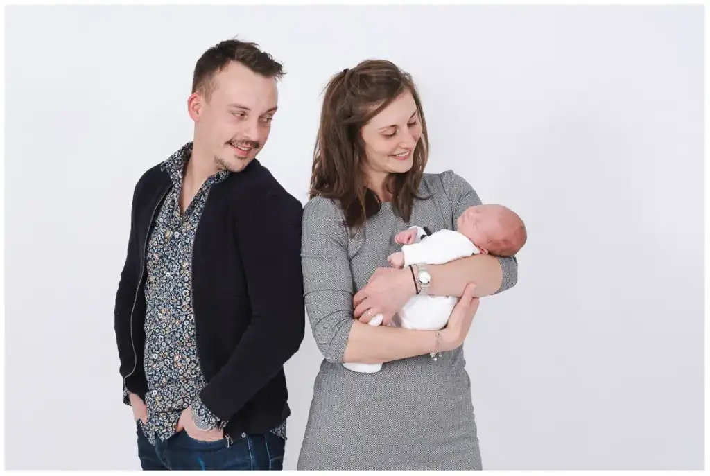 Deux parents regardent leur nouveau-né endormi dans les bras de sa mère devant un fond clair.