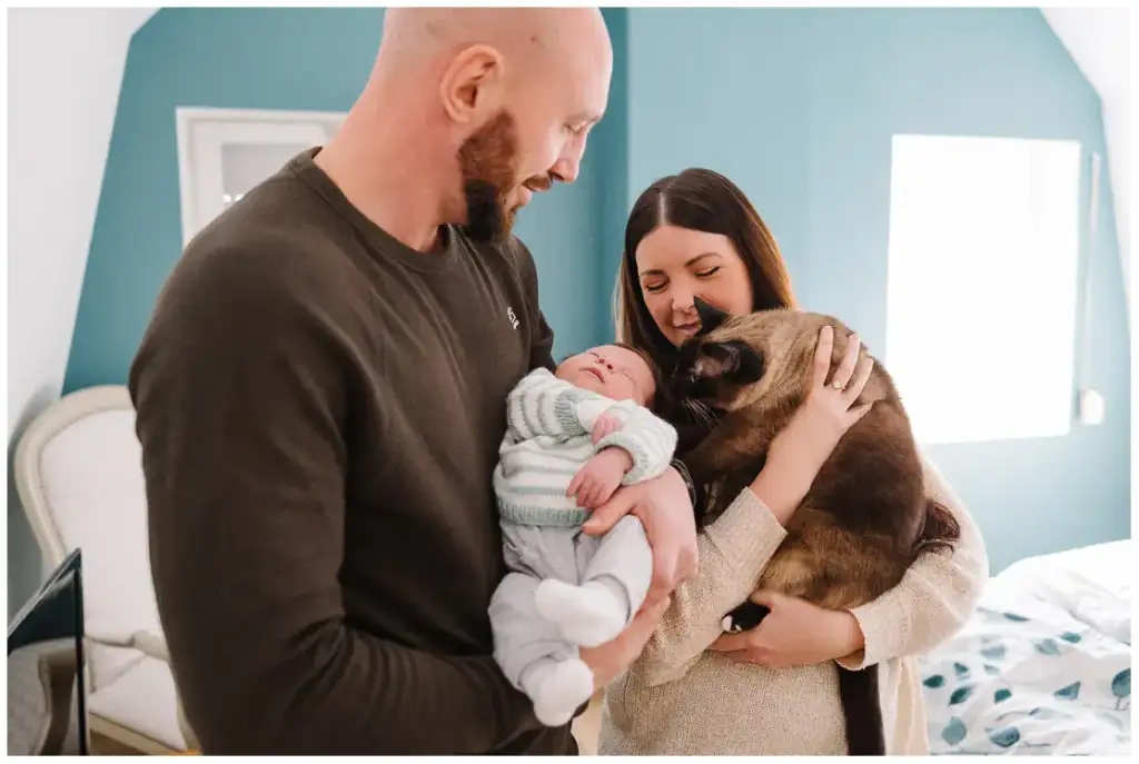 Deux parents tiennent leur nouveau-né et leur chat dans une chambre aux murs bleus.