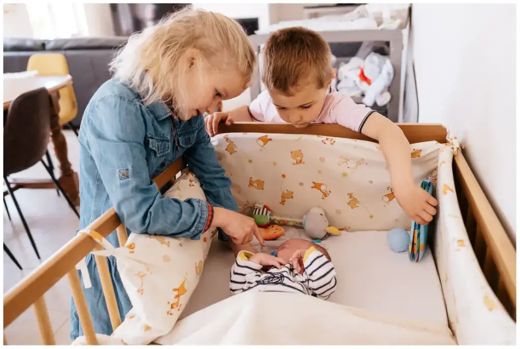 Deux jeunes enfants se penchent au-dessus du lit de leur nouveau-né dans une chambre.