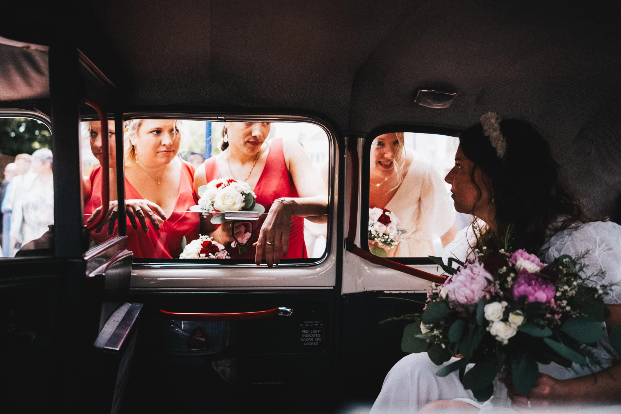 Mariée assise dans une voiture ancienne et entourée de proches tenant leurs bouquets.